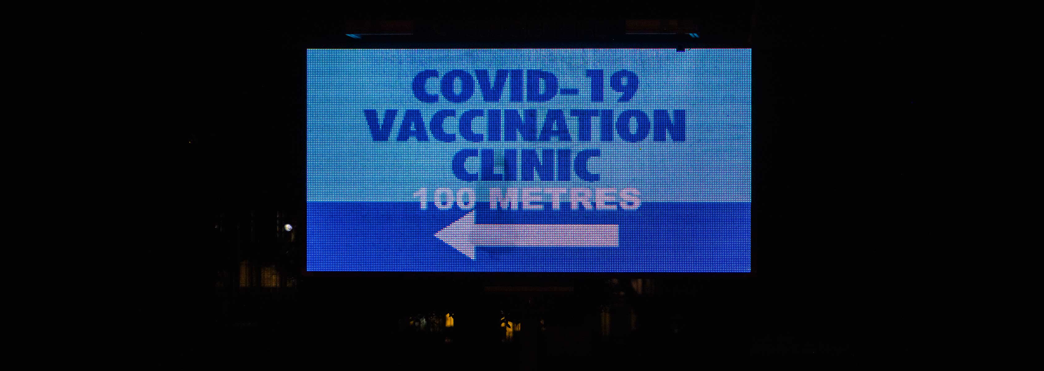 Roadside electronic sign which reads COVID-19 VACCINATION CLINIC 100 METRES with a white arrow pointing to the left. 