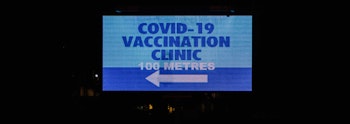 Roadside electronic sign which reads COVID-19 VACCINATION CLINIC 100 METRES with a white arrow pointing to the left. 